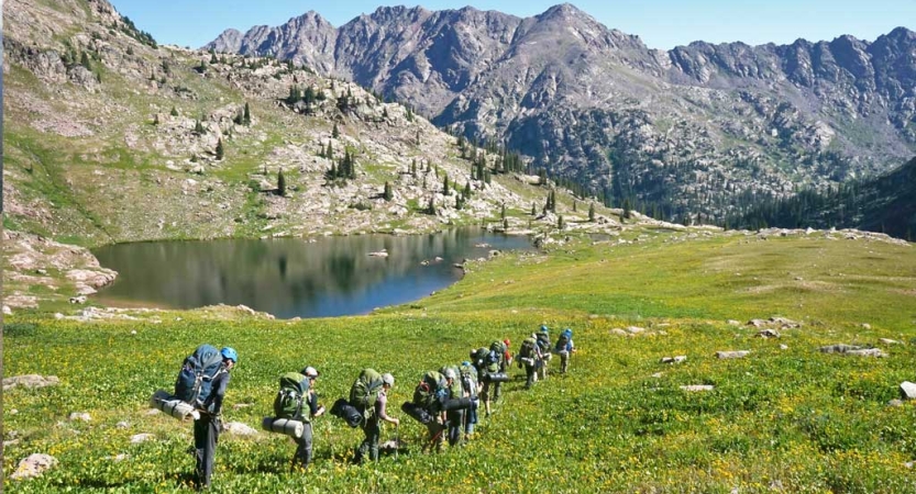 A group of students carrying backpacks hike through a green open area with an alpine lake and mountains in the background
